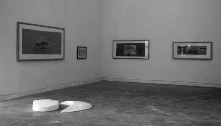 Ein leerer Ausstellungsraum mit Bildern an den Wänden und zwei Skulpturen auf dem Boden.