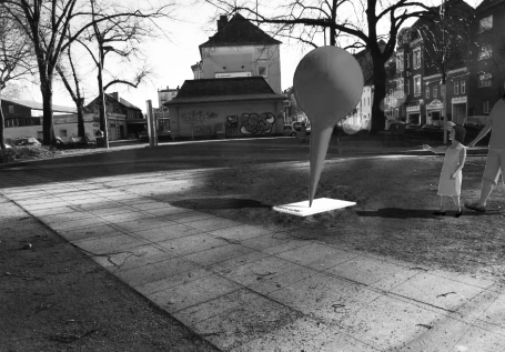 Eine abstrakte Skulptur steht auf einem Platz, umgeben von Bäumen und Gebäuden.